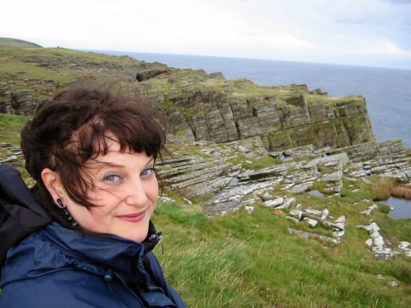 a white woman with dark hair and blue waterproofs looks into the camera. Behind her wide cliffs and open sea