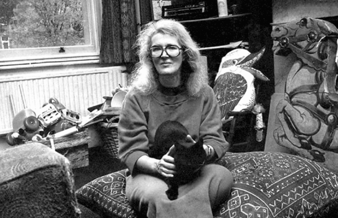 a white woman with long grey hair sits in a living room filled with toys holds a black cat on her lap