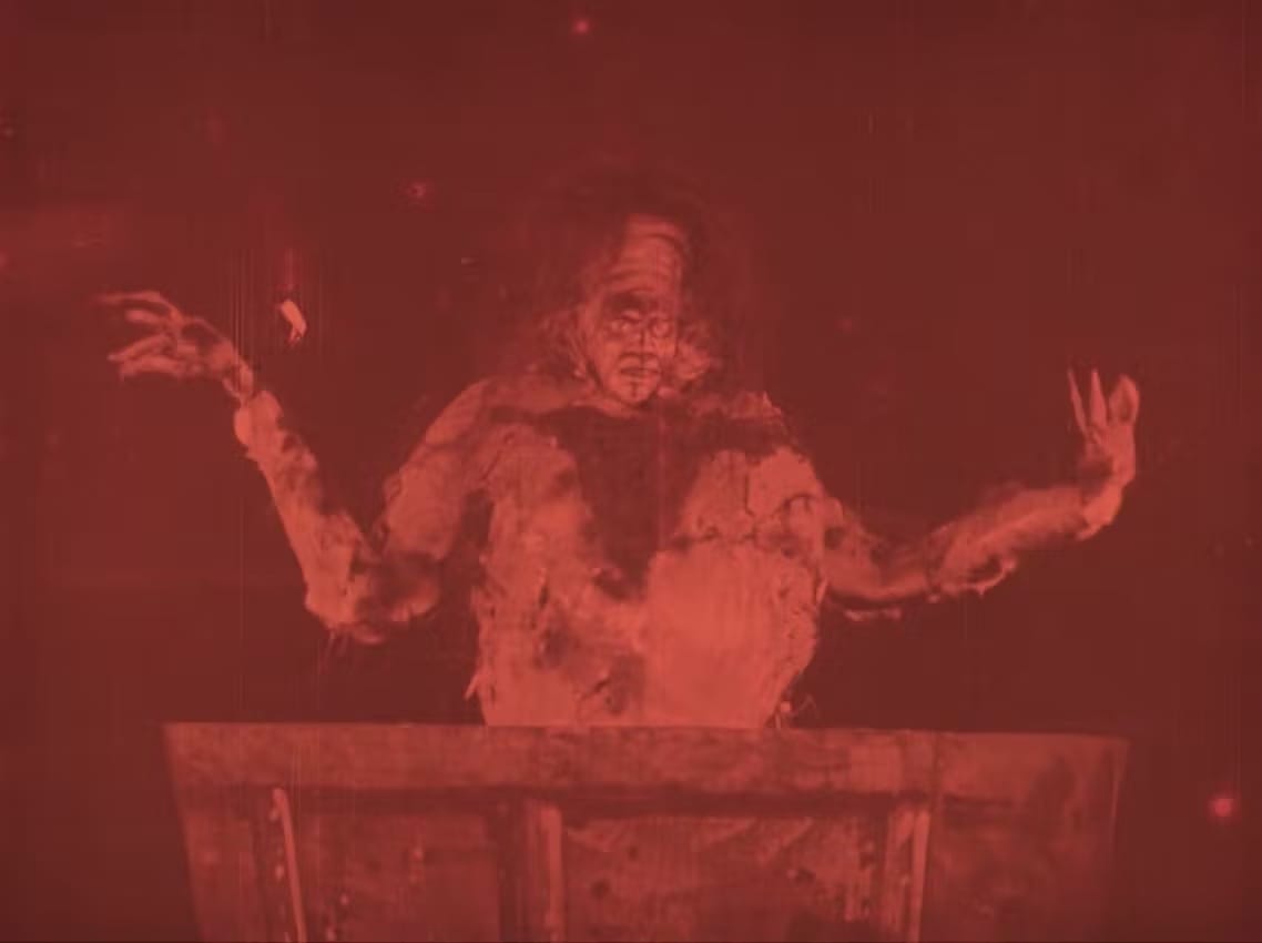 A red toned film still of a grotesque corpse-puppet with piercing eyes